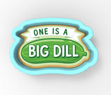 A cookie cutter shaped like a pickle with the text "ONE IS A BIG DILL" printed on it.