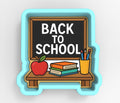 A blue cookie cutter shaped like a chalkboard with the text "BACK TO SCHOOL" written on it, along with various school supplies such as books, pencils, and an apple.
