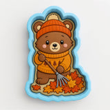 A brown bear wearing a sweater and hat is holding a rake and standing on a pile of autumn leaves.