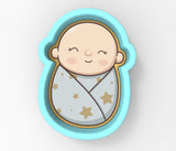 A cartoon image of a baby wrapped in a blanket with stars on it, depicted in a blue and gold cookie cutter.