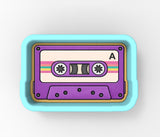 A purple and white cassette tape with a pink and yellow stripe, featuring a white circle with a black dot and a black dot on the left side.