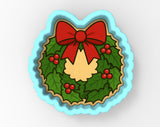 A wreath-shaped cookie cutter with a red bow and holly leaves, berries, and a yellow center, set against a light blue background.