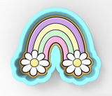 A colorful, cartoon-style cookie cutter shaped like a rainbow with two white daisy flowers on either side.