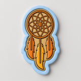 A cookie cutter in the shape of a dreamcatcher, with a brown wooden frame and three orange feathers.