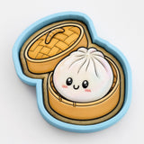 A cookie cutter in the shape of a smiling dumpling, with a yellow lid and a brown interior, is depicted in the image.