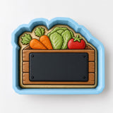 A blue cookie cutter shaped like a wooden crate, with a blackboard-like surface inside. The cutter is filled with various vegetables, including carrots, lettuce, and tomatoes.