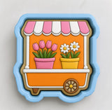 A cookie cutter in the shape of a flower shop window, featuring pink tulips and white daisies in pots.