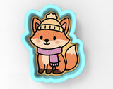 A cartoon image of a fox wearing a hat and scarf, sitting in a blue cookie cutter shape.