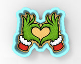 A cookie cutter in the shape of a heart, with two green hands wearing Santa hats on either side of the heart.