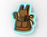 A cookie cutter in the shape of a brown apron with pockets, holding various hair styling tools such as scissors, combs, and a hairbrush.