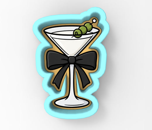 A cookie cutter shaped like a martini glass with a black bow around it, and green olives on top.