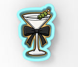 A cookie cutter shaped like a martini glass with a black bow around it, and green olives on top.