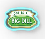 A cookie cutter shaped like a pickle with the text "ONE IS A BIG DILL" printed on it.