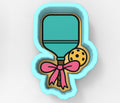 A cookie cutter in the shape of a paddle with a pink bow and a yellow ball.