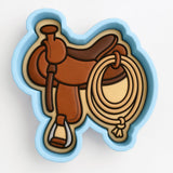 A blue cookie cutter shaped like a brown leather saddle with a rope and a stirrup.