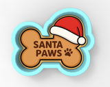 A cookie cutter shaped like a bone with a Santa hat on top and the text "SANTA PAWS" written on it.