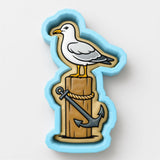 A seagull standing on a wooden post with an anchor, depicted in a cookie cutter-like shape.