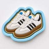 A pair of white and black Adidas sneakers with blue accents, displayed in a blue and white cookie cutter-shaped container.