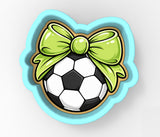 A cookie cutter in the shape of a soccer ball with a green bow on top.