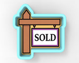 a 3D rendering of a "SOLD" sign, which is a rectangular sign with the word "SOLD" written in bold black letters on a white background. The sign is attached to a wooden post, and the overall design is simple and straightforward.