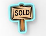 A blue cookie cutter shaped like a "SOLD" sign, with the word "SOLD" written in bold black letters on a light brown background.