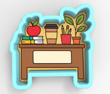 A blue cookie cutter shaped like a desk with various school supplies and a plant on it.