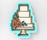 A cookie cutter in the shape of a cake, with a white cake layer and three orange roses on top.