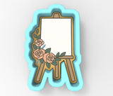 A 3D rendering of a white canvas on a wooden easel, decorated with pink roses and green leaves.